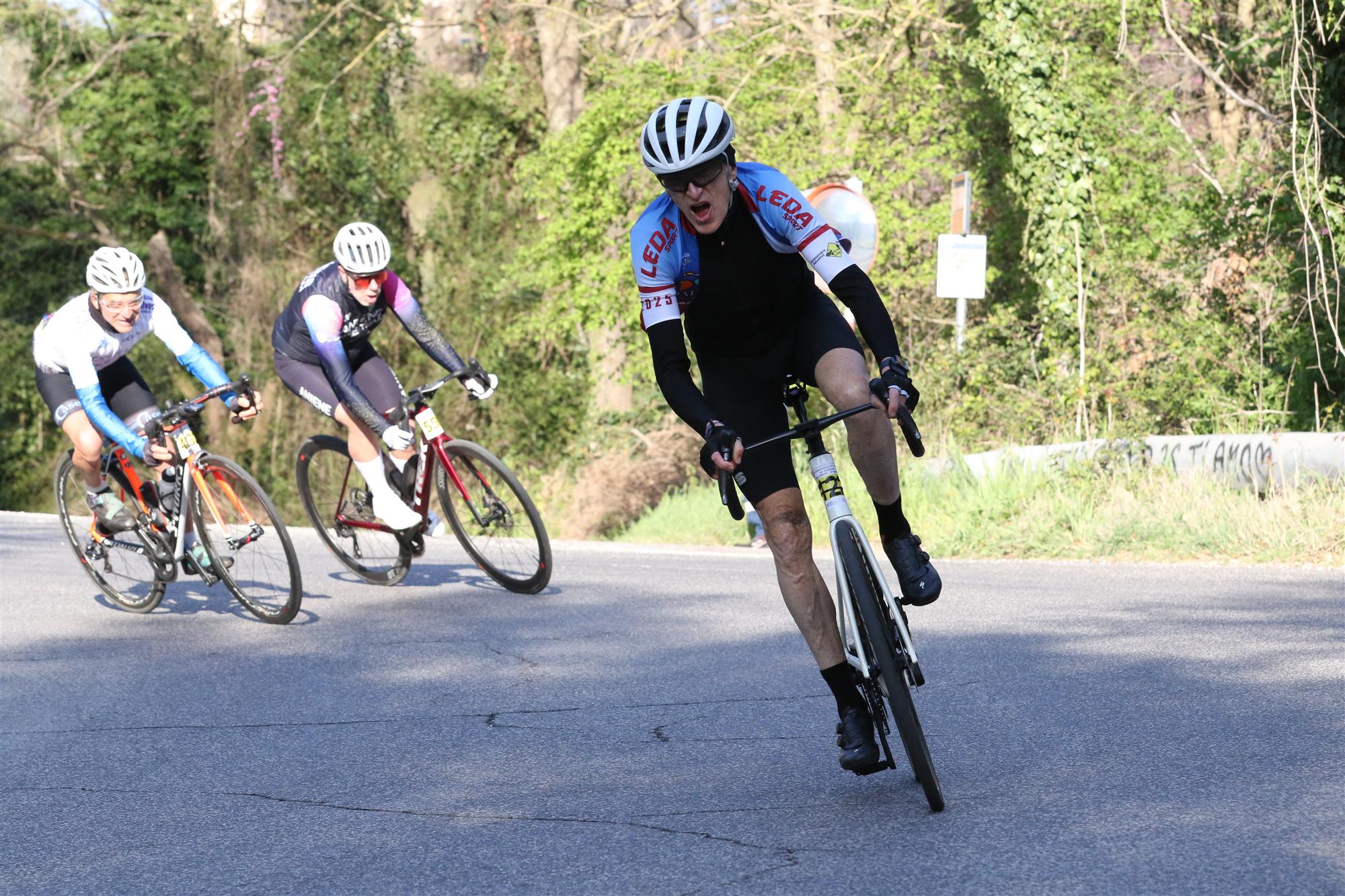 Tre ciclisti in gara su una strada fra gli alberi